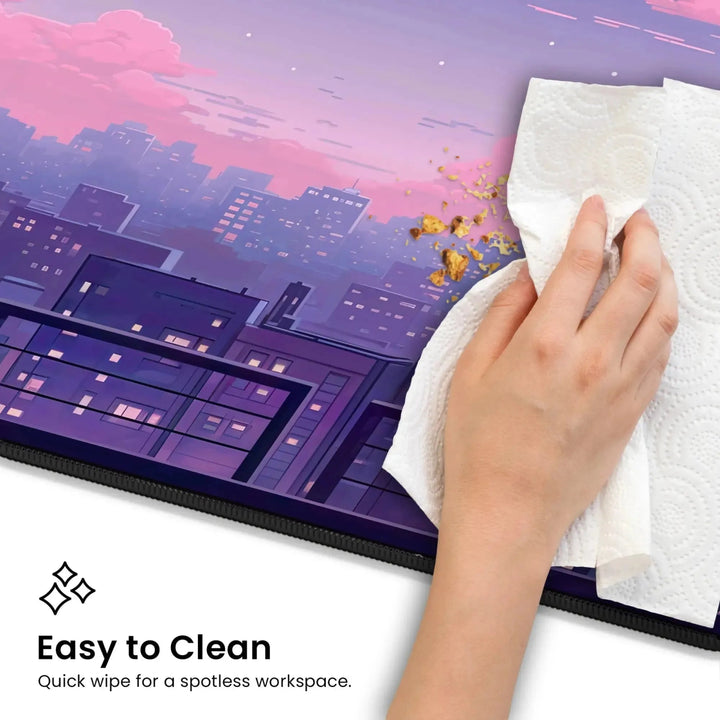 A hand using a paper towel to clean a spill on the Kawaii Desk Mat – Lofi Vaporwave Pixel Art Anime Mouse Pad for Gaming & Work, featuring a cityscape illustration at dusk. The text reads, "Easy to Clean - Quick wipe for a spotless workspace." The background has a purple and pink sky with illuminated buildings.