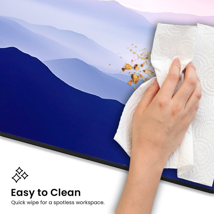 Home Decor - A hand uses a paper towel to wipe crumbs off a surface with a blue and purple abstract mountain design. The text in the image reads, "Easy to Clean - Quick wipe for a spotless workspace." This Mountain Desk Mat – Large Nature Mouse Pad for Office or Gaming Setup not only provides scenic views but also ensures hassle - free maintenance. - Kaladyn Designs
