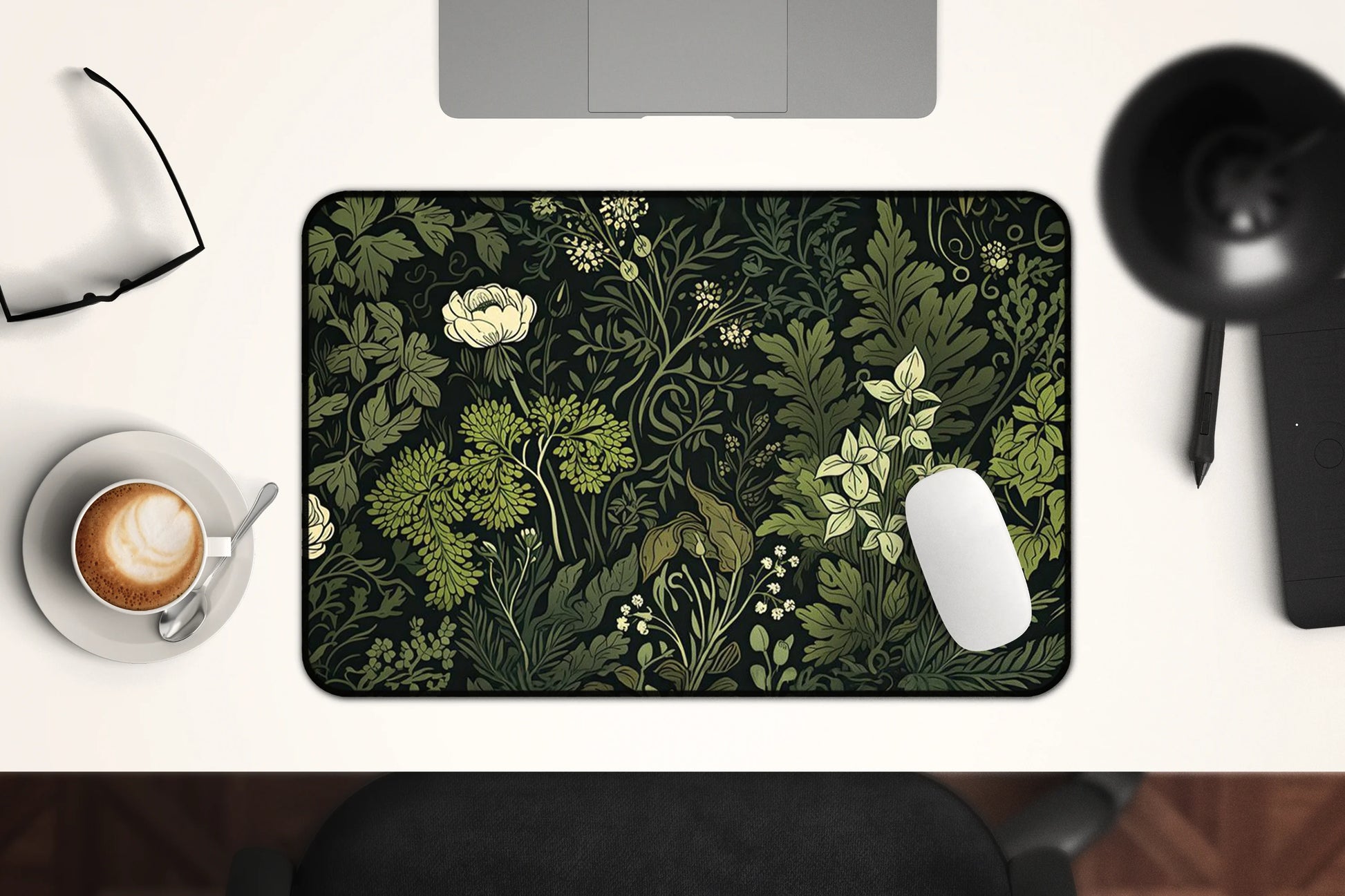 Overhead view of a desk setup featuring a dark green floral-themed desk mat with botanical illustrations. Includes a monitor, keyboard, mouse, coffee cup, and glasses on a white desk surface.