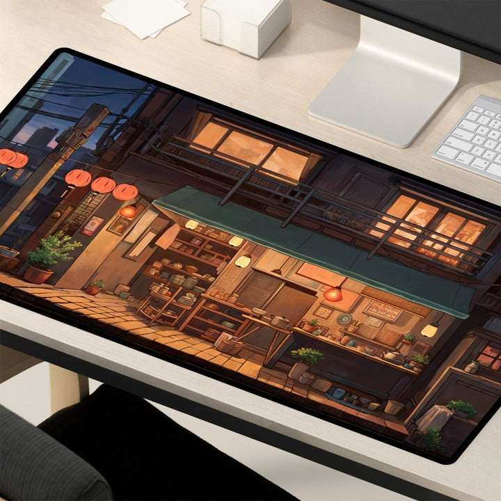 Home Decor - A desk featuring a computer monitor displaying artwork of a cozy, illuminated Japanese street scene at dusk. The street has a small café with hanging lanterns, a green awning, and a warm, inviting atmosphere. Nearby are a keyboard and mouse atop an Anime Shop Desk Mat - Lofi Aesthetic, Large Mouse Pad, perfect for aesthetic desk decor and gaming setups, along with office supplies. - Kaladyn Designs