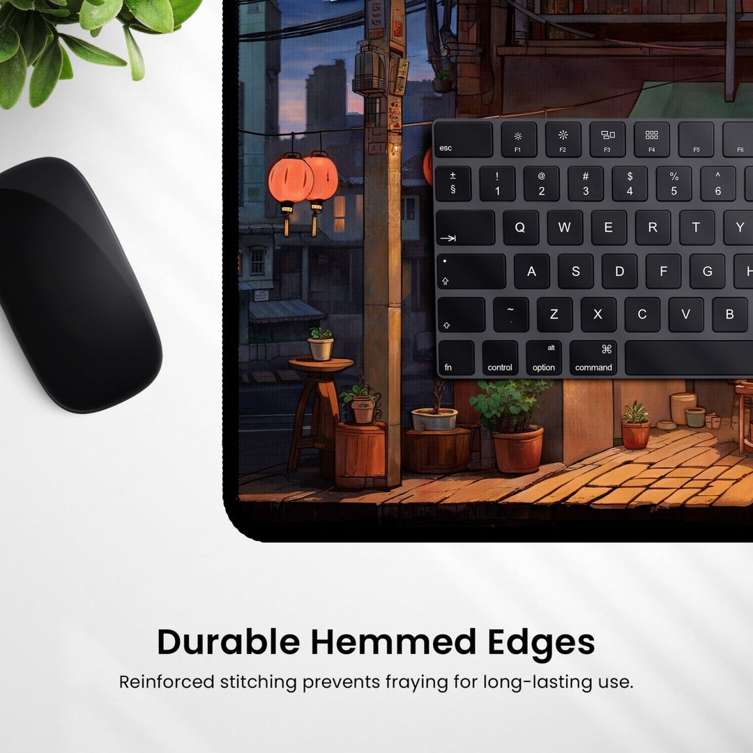 Home Decor - An Anime Shop Desk Mat, also serving as a gaming mouse pad, showcases a portion of a keyboard and mouse on its surface. The lofi aesthetic design depicts a cozy street scene adorned with potted plants and lanterns. Text at the bottom reads, "Durable Hemmed Edges: Reinforced stitching prevents fraying for long - lasting use. - Kaladyn Designs