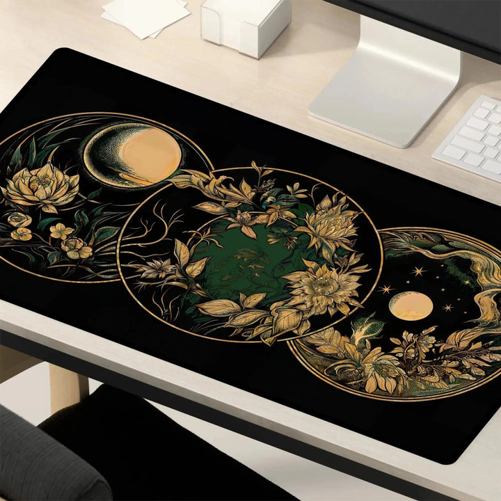 A desk setup featuring the Art Nouveau Celestial Moon Desk Mat – Witch Aesthetic Gaming Mouse Pad. The large mat is adorned with intricate gold and green floral and celestial designs, including stylized representations of a sun, moon, and stars amid leaves and flowers, evoking an Art Nouveau charm. A computer monitor and keyboard are also visible.