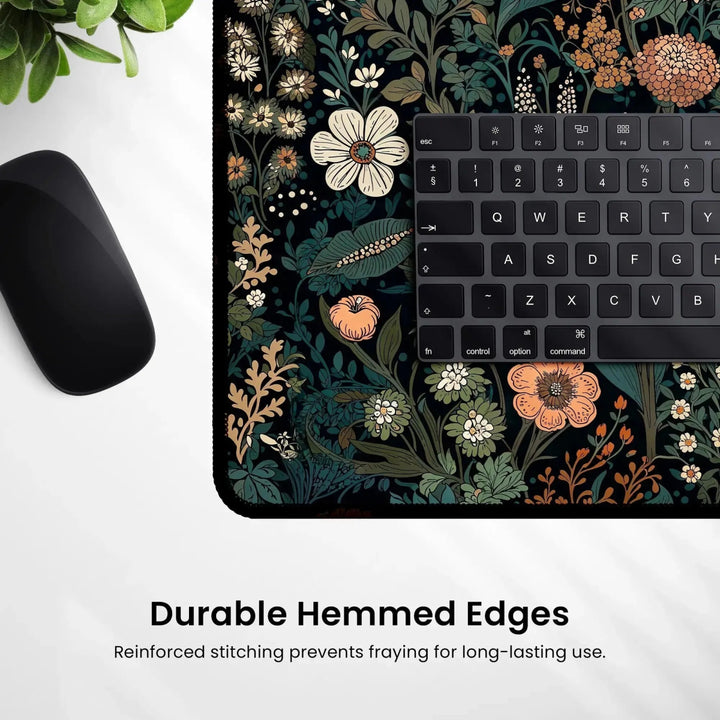 A close-up of the Dark Cottagecore Botanical Desk Mat with a vibrant floral pattern and a section of a black keyboard on it. The desk mat features durable hemmed edges, exuding a distinct dark cottagecore aesthetic. A computer mouse rests on one side, and a green plant is partially visible in the top corner. Text reads "Durable Hemmed Edges.