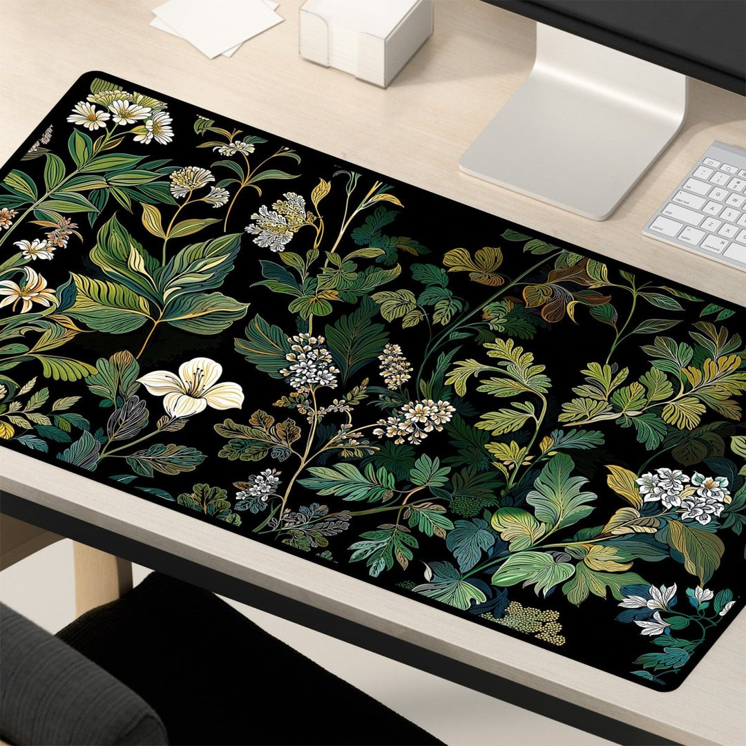 Home Decor - A desk is seen with a keyboard, monitor, and a large Elegant Botanical Black Floral Desk Mat featuring green and white leaves and flowers on a black background. A chair is partially visible in the foreground. - Kaladyn Designs