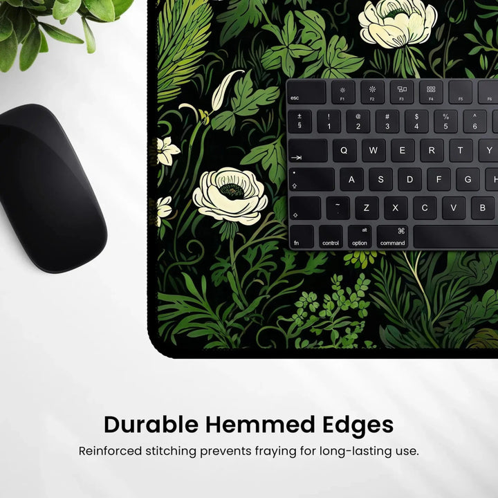 Home Decor - A Floral Sage Green Botanical Desk Mat showcasing white flowers and green leaves, paired with a black computer keyboard and mouse. Text at the bottom reads "Durable Hemmed Edges: Reinforced stitching prevents fraying for long - lasting use." A plant is partially visible in the top left corner. - Kaladyn Designs