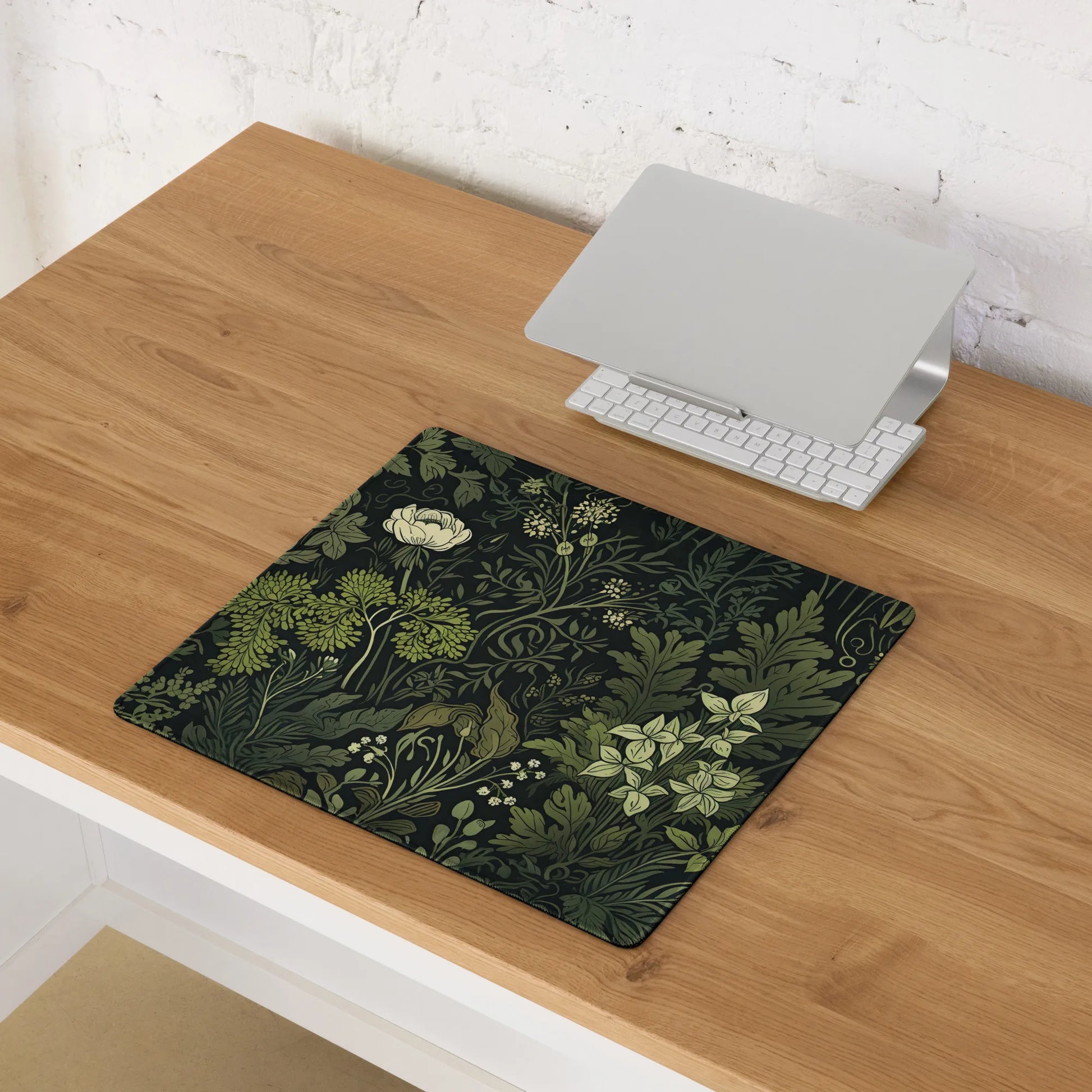 Overhead view of a compact dark green floral-themed desk mat with intricate botanical illustrations on a wooden desk, featuring a laptop and keyboard.