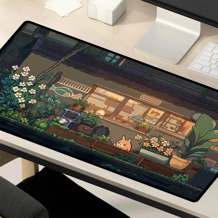 A minimalist desk enhanced by a colorful, animated **Kawaii Desk Mat – Cute Japanese Anime Design Non-Slip Mouse Pad for Gamers** that features a charming garden scene. The mat showcases a café surrounded by plants, flowers, and adorable animals, including a small cat enjoying the view. A computer monitor and keyboard rest nearby on the desk.