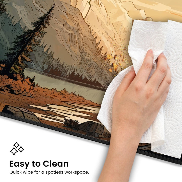 Home Decor - A hand uses a paper towel to wipe the surface of the durable "Mountain Desk Mat – Large Nature Desk Mat for Gaming & Office, Scenic Forest and Valley Design." The image includes text that reads, "Easy to Clean: Quick wipe for a spotless workspace." This nature - inspired decor brings tranquility to your workstation. - Kaladyn Designs