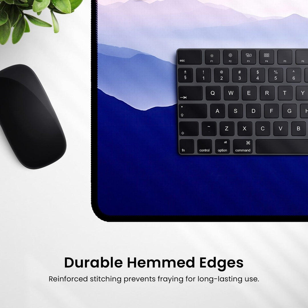 Home Decor - A black keyboard rests on a Mountain Desk Mat – Large Nature Mouse Pad for Office or Gaming Setup, featuring a scenic mountain design with durable hemmed edges. A black computer mouse and a small green plant are also partially visible in the image. Text reads: "Durable Hemmed Edges - Reinforced stitching prevents fraying for long - lasting use. - Kaladyn Designs