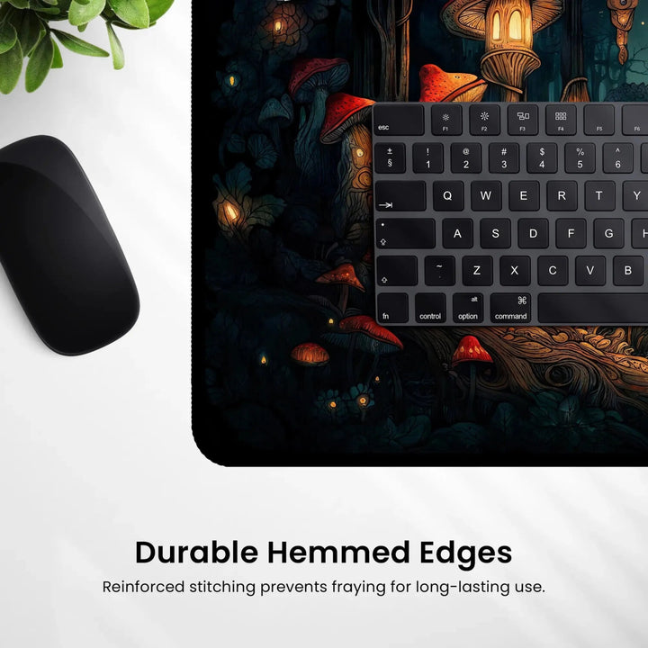 Resting on a dark-themed Mousepad, the computer keyboard and mouse create an ideal gaming setup with a fantasy forest design featuring mushrooms and glowing lanterns. The Mushroom Forest Desk Mat – Dark Cottagecore Witchy Gaming Mouse Pad & Tarot Mat is equipped with durable hemmed edges to prevent fraying, as noted below the image.